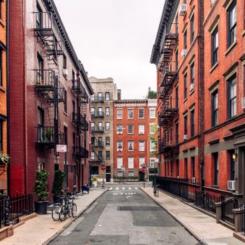 Bicycles rest against iron fences on a narrow street lined with red-brick buildings, with fire escapes and a "STOP" marked on the road.
