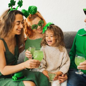 People dressed in green are toasting with green drinks, celebrating joyfully; they wear shamrock-themed accessories indoors.