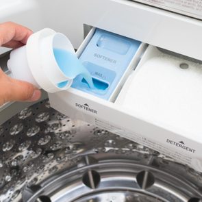 Hand pouring blue liquid into a washing machine's softener compartment, next to a detergent tray with white powder, inside a stainless drum.