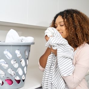 Woman smelling clean laundry; standing in a laundry room with a gray basket of clothes on a white counter.