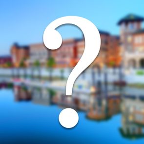 A large white question mark overlays a blurred image of colorful buildings reflecting on a calm water surface in a sunny setting.