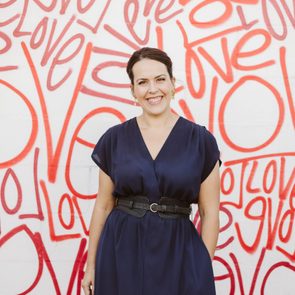 A person stands smiling, wearing a navy dress with a belt, against a white wall covered in red "love" graffiti text.