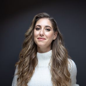A person with long, wavy hair, wearing a white sweater, smiles gently against a dark background.