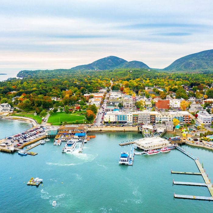 Aerial view shows a harbor lined with vibrant trees and buildings, nestled between a calm bay and distant green mountains.