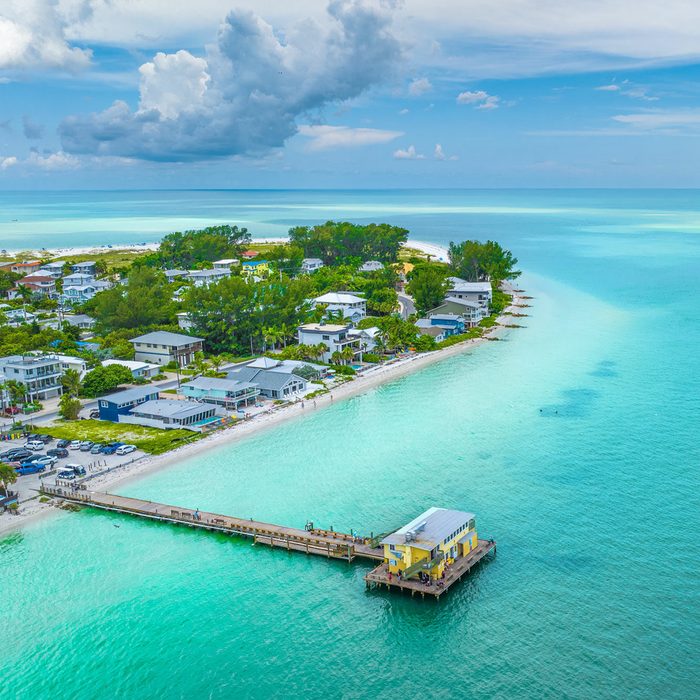 Pier extends over turquoise water, bordered by beachfront houses and lush trees, under a partly cloudy sky.