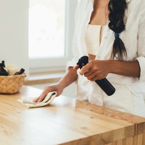 Person cleans a wooden table using a spray bottle and cloth, standing in a bright kitchen next to a basket with bottles.