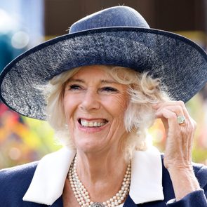 A woman wearing a large blue hat smiles brightly, adjusting her hair, against a vibrant, blurred garden background.
