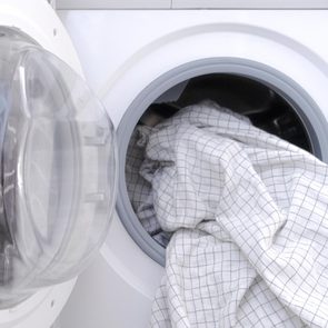 A front-loading washing machine contains a crumpled, white grid-patterned fabric, set in a bright laundry room.