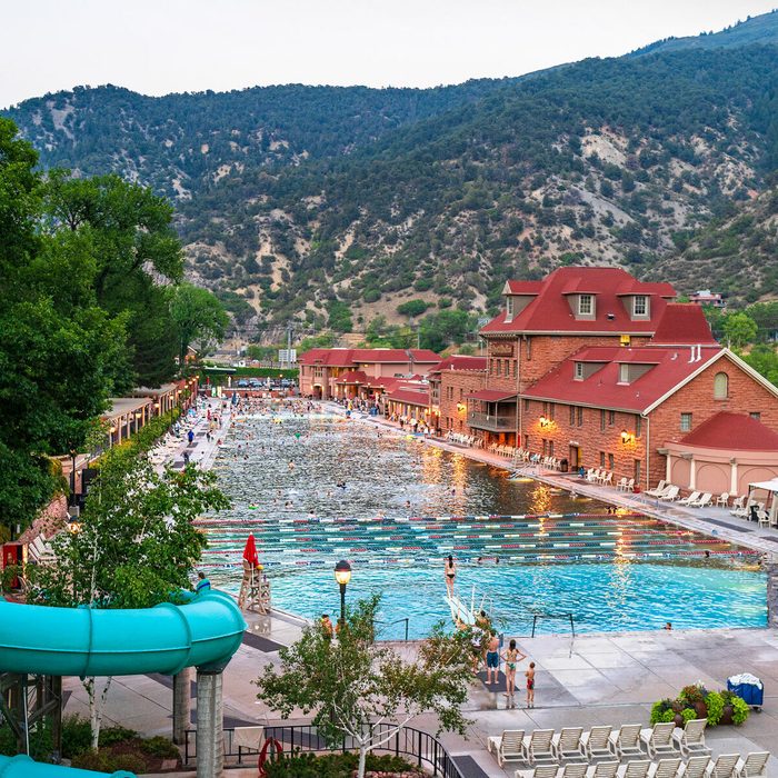 A large outdoor pool with swimmers is surrounded by a red-roofed building and lush trees, set against mountainous terrain.