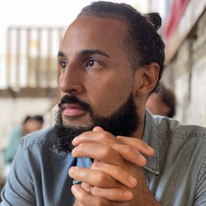 A person with clasped hands gazes thoughtfully, sitting in a casual indoor setting with blurred background elements.