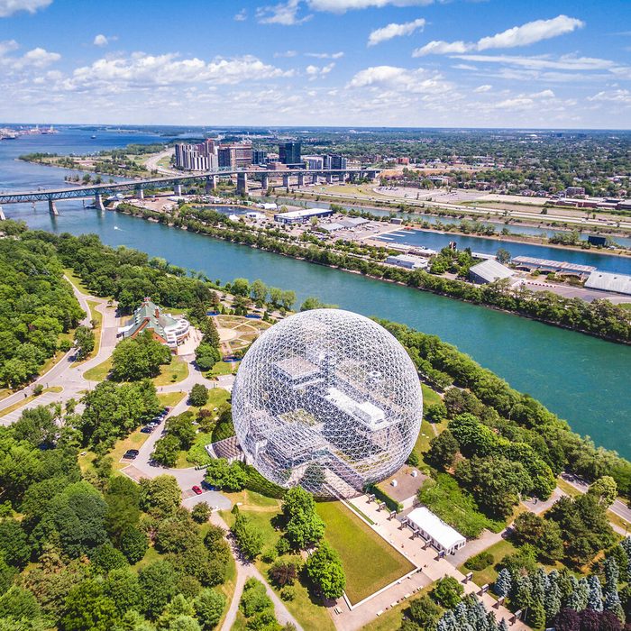 Geodesic dome stands among green trees beside a river, with a bridge and cityscape visible in the background under a blue sky.