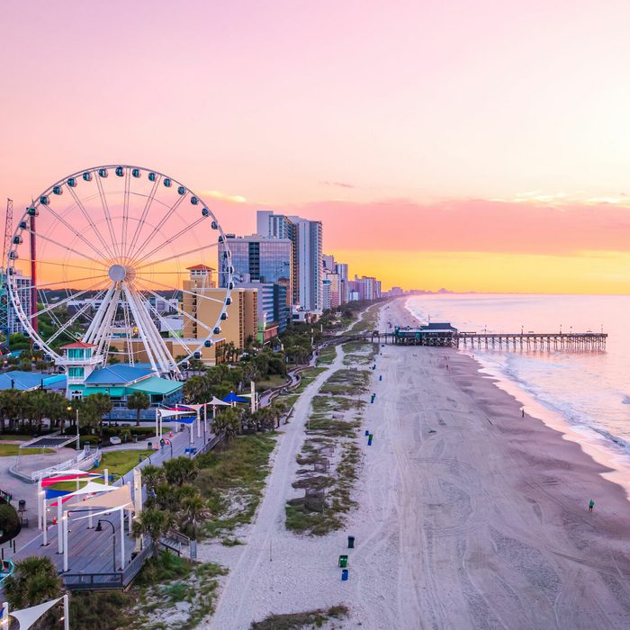 Ferris wheel stands tall beside a beach, glowing under a vibrant sunset sky, with a cityscape and ocean pier in the background.