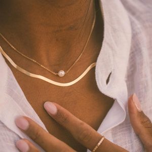 Necklaces adorn neck, highlighting a pearl pendant. Fingers gently touch a white fabric shirt, creating an elegant and minimalist style.