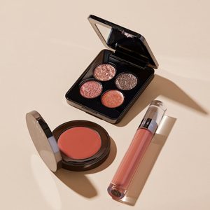 A makeup palette with shimmery eyeshadows sits open on a beige surface, alongside a blush compact and a lip gloss tube.
