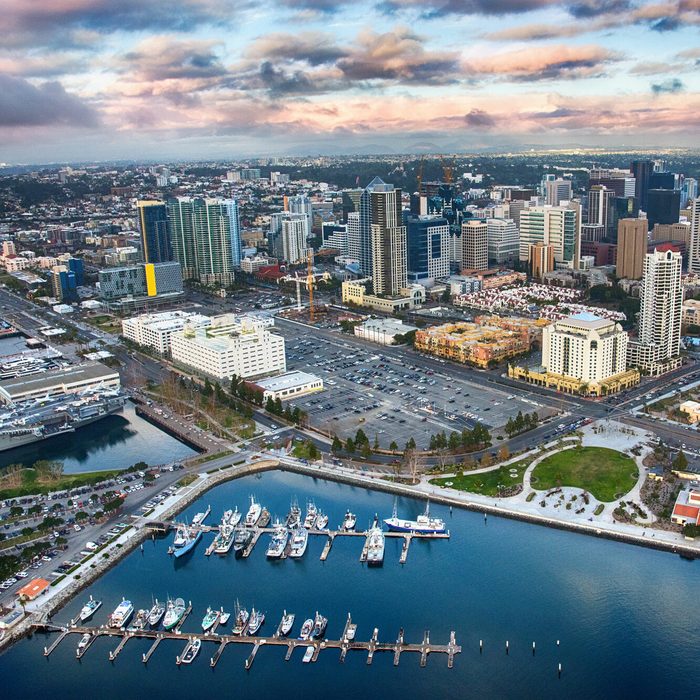 Harbor filled with docked boats, surrounded by urban cityscape under a cloudy sky, buildings and roads bustling, green park visible near waterfront.