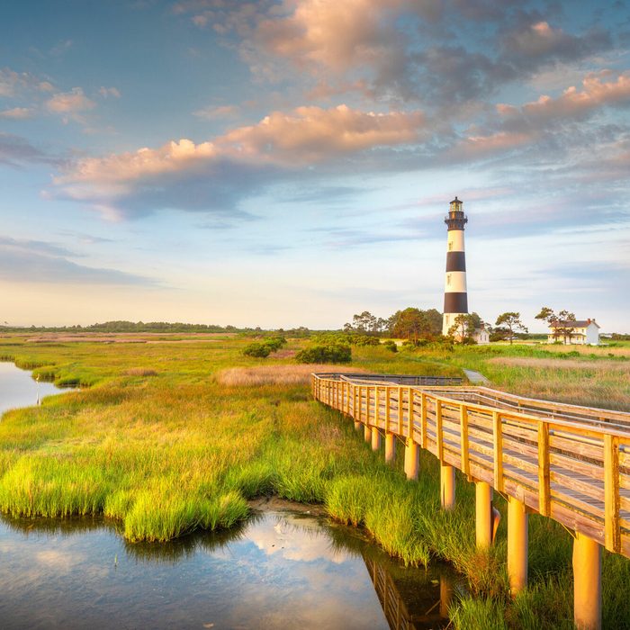 Lighthouse stands tall, overlooking a wooden boardwalk across lush wetlands under a partly cloudy sky at sunset.