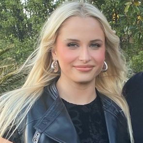 A person is smiling, wearing hoop earrings and a black jacket, standing outdoors with greenery in the background.