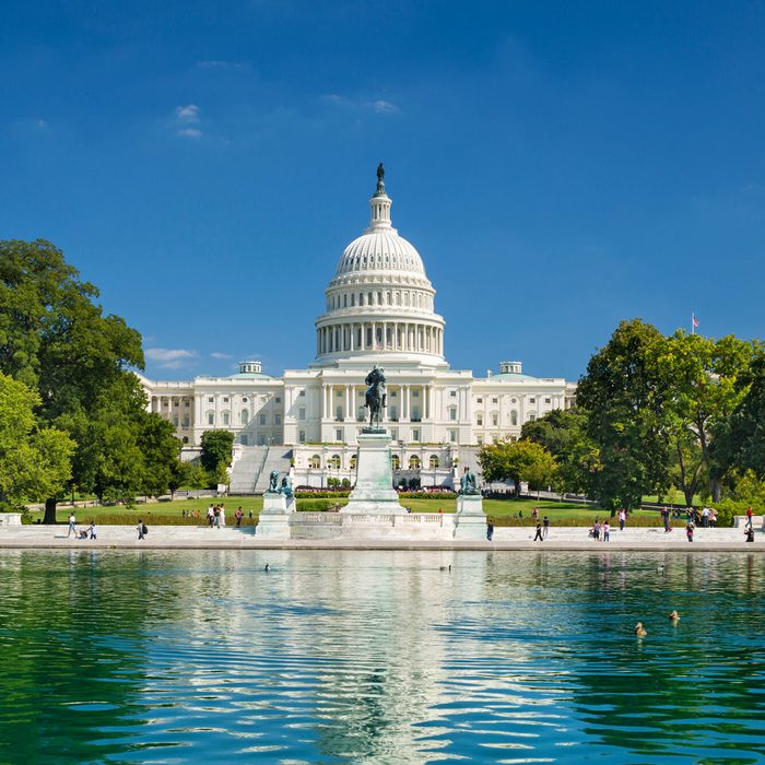 The Capitol building stands tall, surrounded by lush trees and a reflecting pool, with people walking along the pathway.