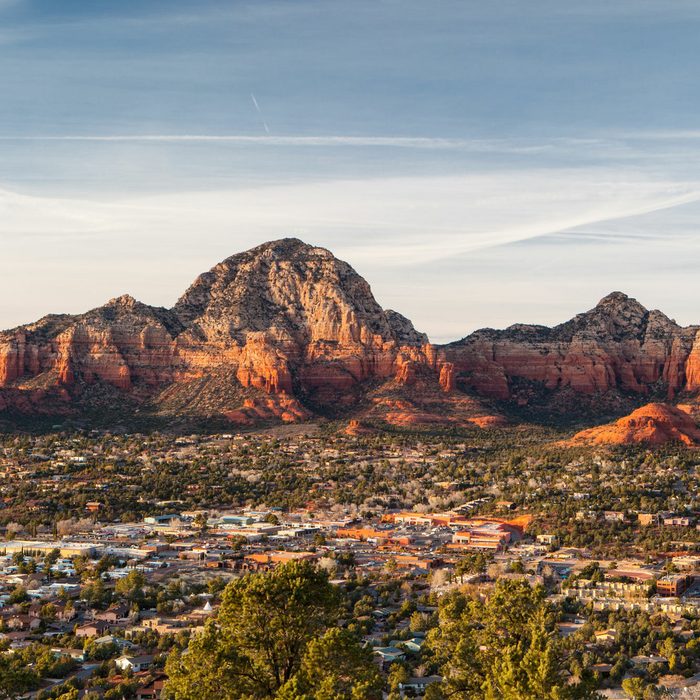 Red rock mountains tower over a sprawling town, nestled in a desert landscape under a vast, clear sky with subtle cloud streaks.