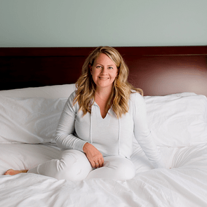 A person sits cross-legged on a bed, wearing white casual clothes, against a dark wooden headboard in a softly lit room.