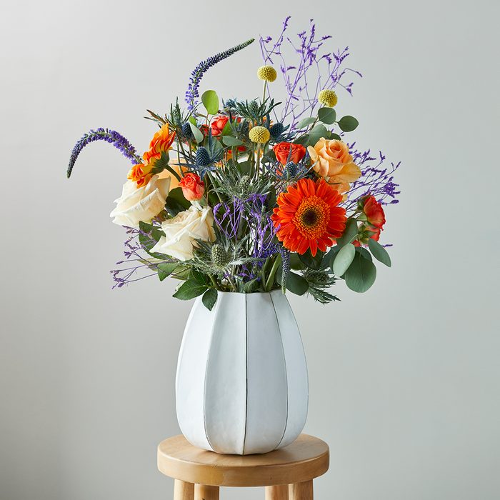 A white vase holds a vibrant floral arrangement on a wooden stool against a plain, light-colored background.