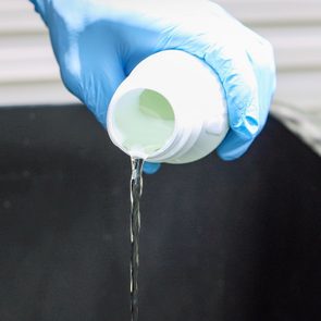 Gloved hand pours clear liquid from a white bottle into a dark container in a bright, outdoor setting.
