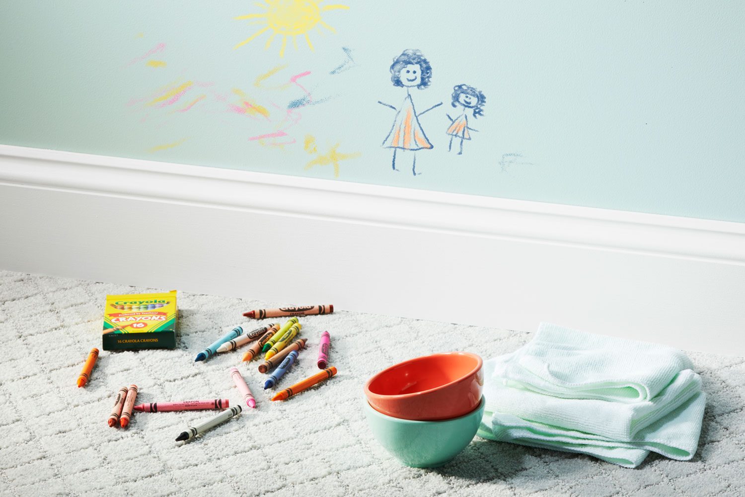 Crayons scattered on carpet, drawing on wall depicting figures and sun, with bowls and cloth nearby in a room.