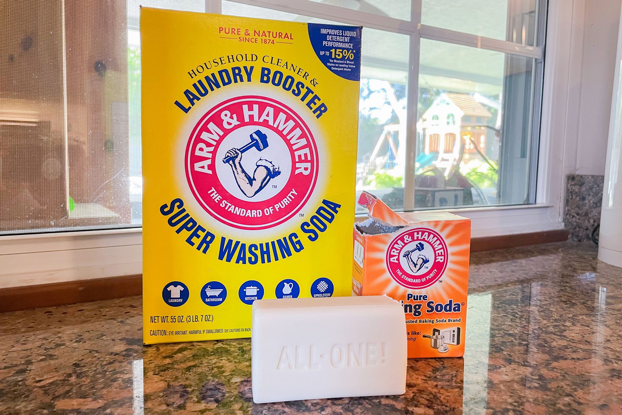 Washing soda box stands beside baking soda box and soap bar on a kitchen counter near a window. Soap reads "ALL-ONE!"