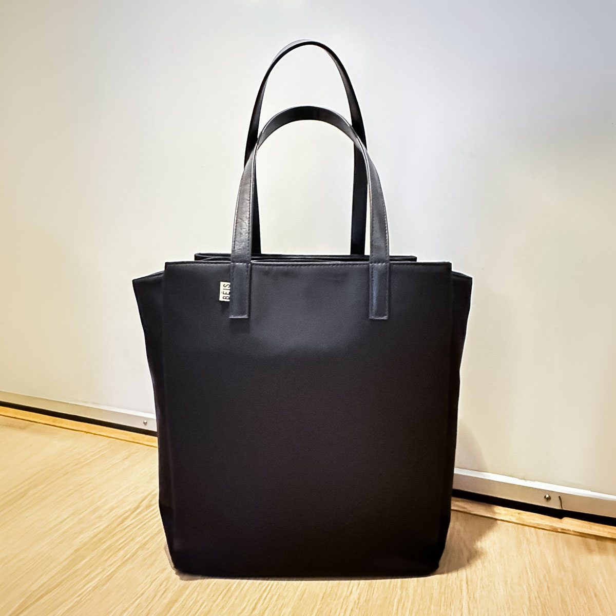 A black tote bag stands against a plain indoor background, featuring two handles and the logo "BEIS" on it.