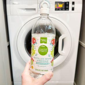 A bottle of distilled white vinegar is held in front of a washing machine in a laundry room.