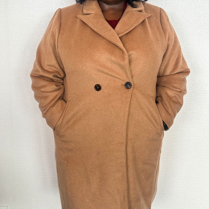 A person stands wearing a tan coat with hands in pockets, against a light textured wall.