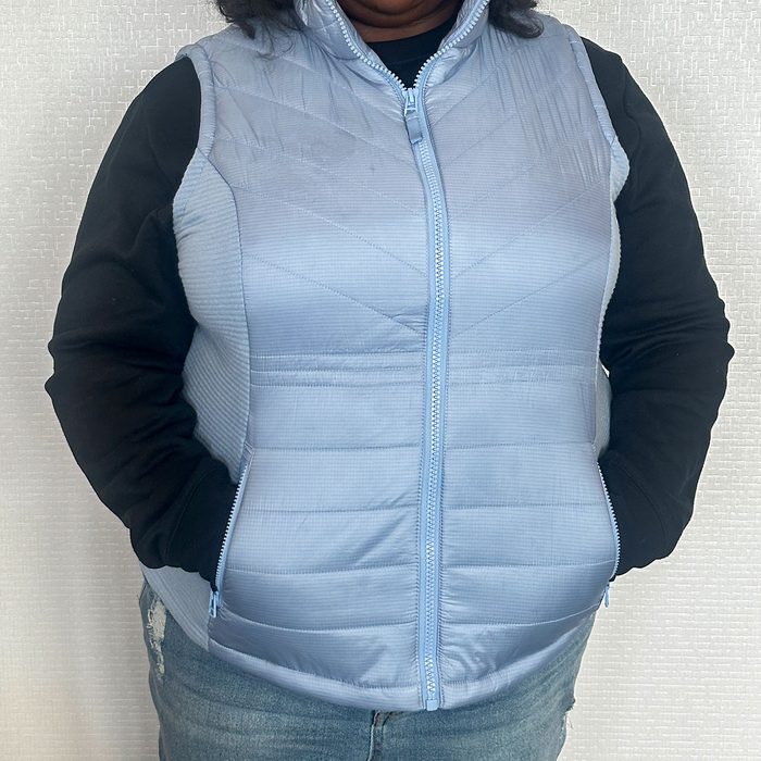 A person wears a light blue, quilted vest over a black long sleeve, standing against a textured light background.
