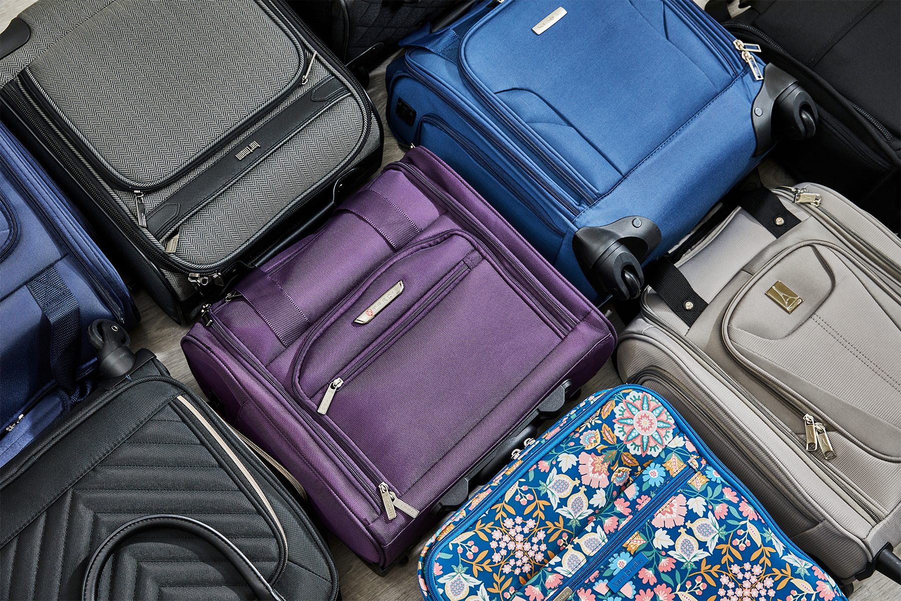 Suitcases are arranged on a floor, showcasing various colors and patterns, including purple, gray, and floral designs, in a crowded storage context.