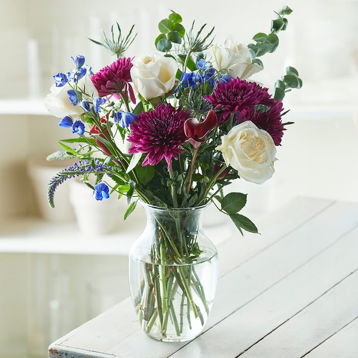 Flowers sit in a clear vase, showcasing white roses, purple and blue blooms on a light-colored wooden table in a softly lit room.