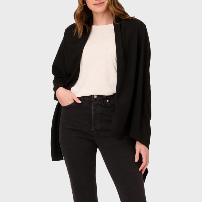 A person wearing a black shawl over a white shirt and black jeans, standing against a light gray background.
