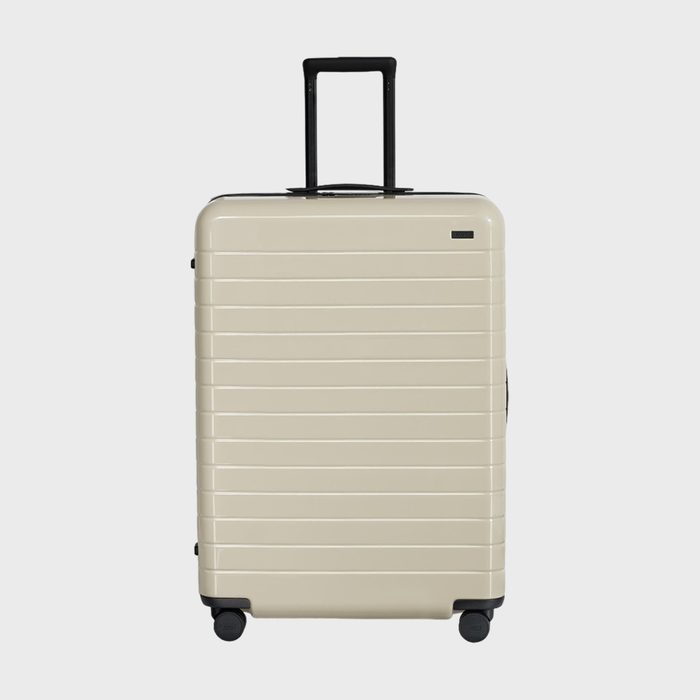 A beige suitcase stands upright, handle extended, on a white background. Four wheels are visible at its base.