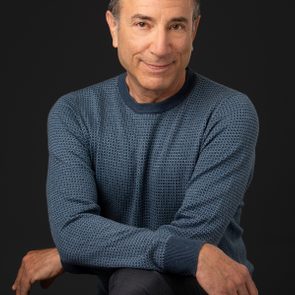 Smiling person sitting on a stool, wearing a blue patterned sweater and dark pants against a dark background.