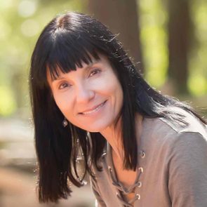 A woman smiles gently, wearing a lace-up shirt, with long dark hair, set against a blurred, sunlit forest background.