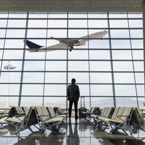 A person stands with luggage, watching a plane take off through large airport windows, amidst rows of empty seats.