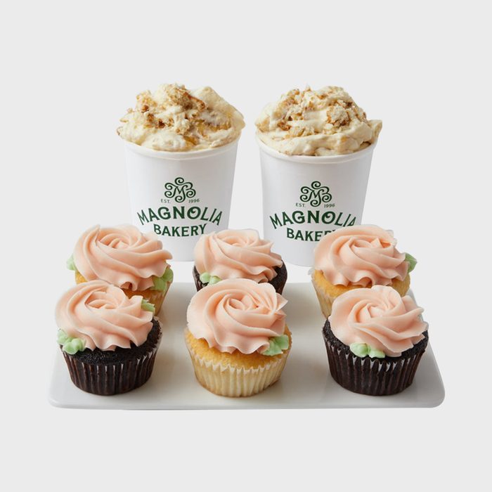 Cupcakes with pink frosting sit on a tray, accompanied by two white cups of Magnolia Bakery pudding with crumb topping.