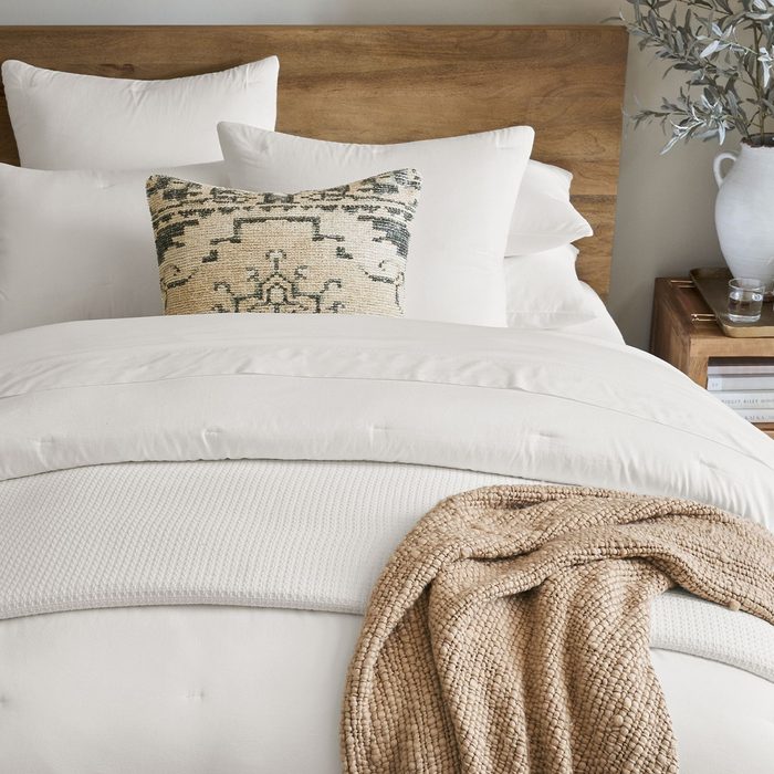 Bed displays layered white bedding with a patterned throw pillow, set in a wooden headboard. A side table with books and vase is nearby.