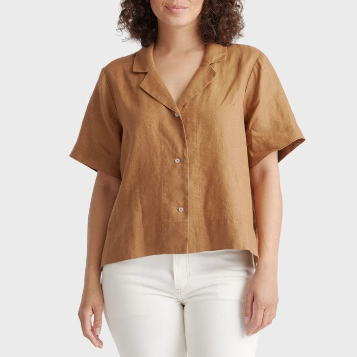 A person stands wearing a brown, short-sleeved button-up shirt and white pants against a plain background.