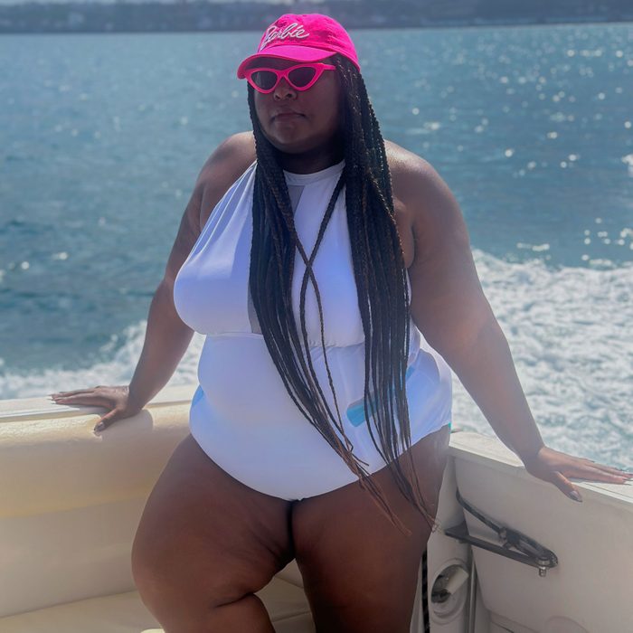Person poses on a boat, wearing a pink hat and sunglasses, with a white swimsuit. Background features a sunny, expansive ocean view.