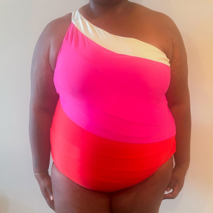 Person wears vibrant, one-shoulder swimsuit with pink and red color blocks, standing against a plain light background.