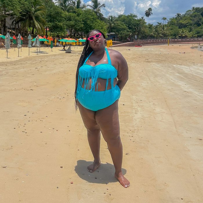 Person stands confidently on a sandy beach wearing a turquoise swimsuit and pink sunglasses. Colorful umbrellas and lush trees are in the background.