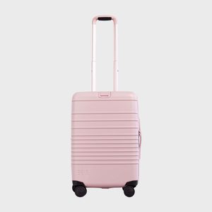 Pink suitcase stands upright, handle extended, on four wheels. Simple design with horizontal grooves. Background is plain gray. 