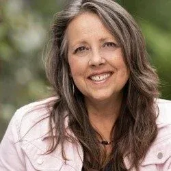 A person smiles warmly, wearing a light jacket, with long hair, set against a blurred green outdoor background.