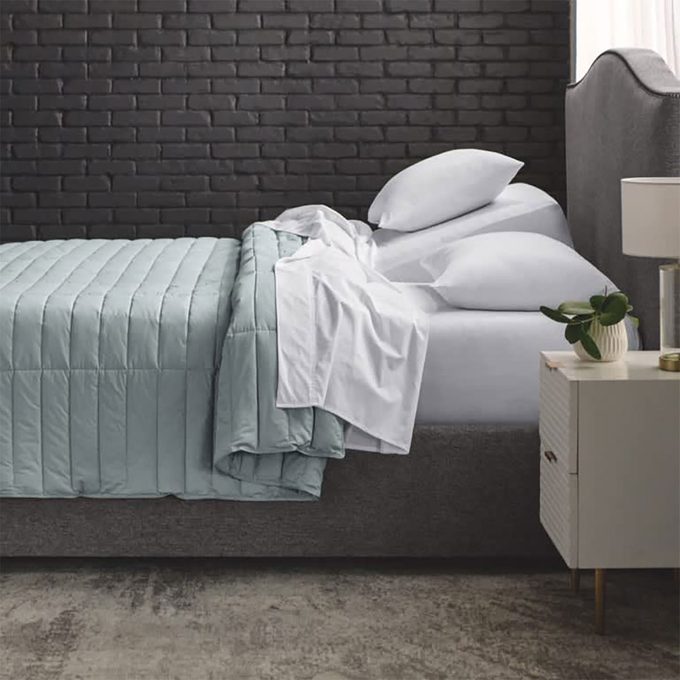 A neatly made bed with pillows sits against a dark brick wall, adjacent to a nightstand holding a lamp and plant.
