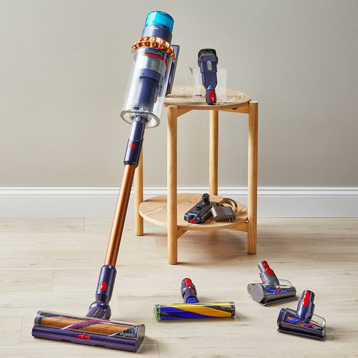 A vacuum cleaner leans against a wall; several attachments rest on a wooden table and floor in a minimalist room.