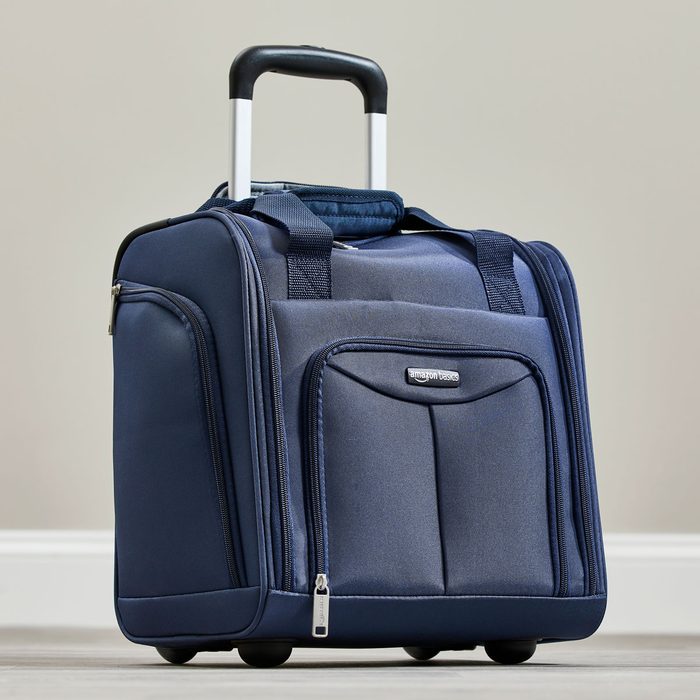 A blue rolling suitcase stands upright on a wooden floor, featuring a telescopic handle and multiple zippered compartments in a neutral setting.